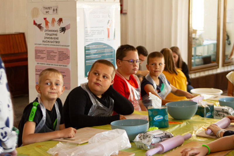 Kinderen zitten aan een tafel tijdens een activiteit in Oekraïne, met kleurrijke materialen en posters op de achtergrond, illustrerend hulp en educatie voor kinderen in conflictgebieden.