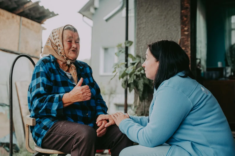 Een jonge vrouw biedt steun en troost aan een oudere vrouw die verdrietig is, in een huiselijke omgeving, onder het thema 'Sponsor het leven' van Dorcas.