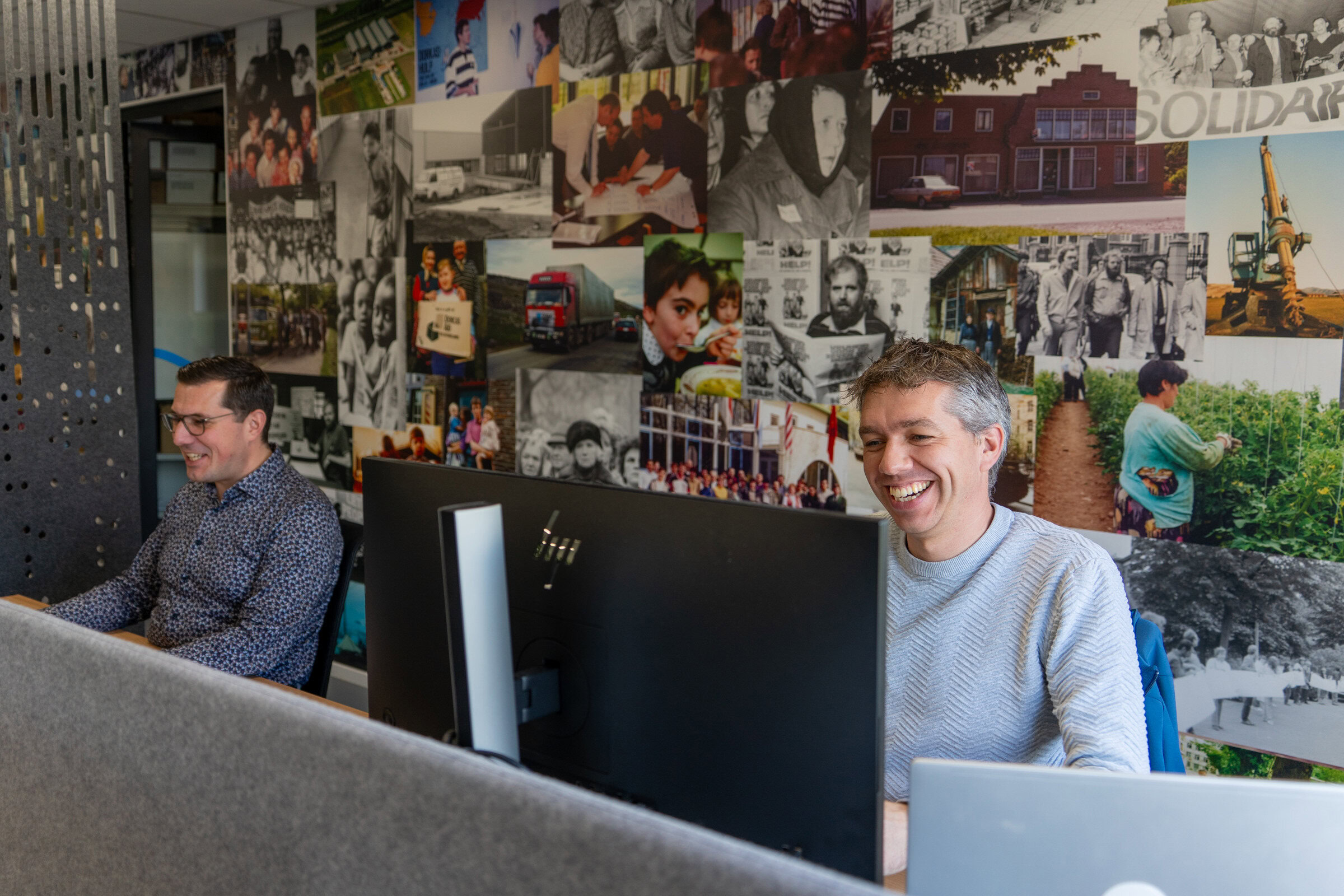 Twee jonge mannen werken blij in een kantoorruimte met een muur vol foto's en posters, passend bij de vacaturepagina van Dorcas.