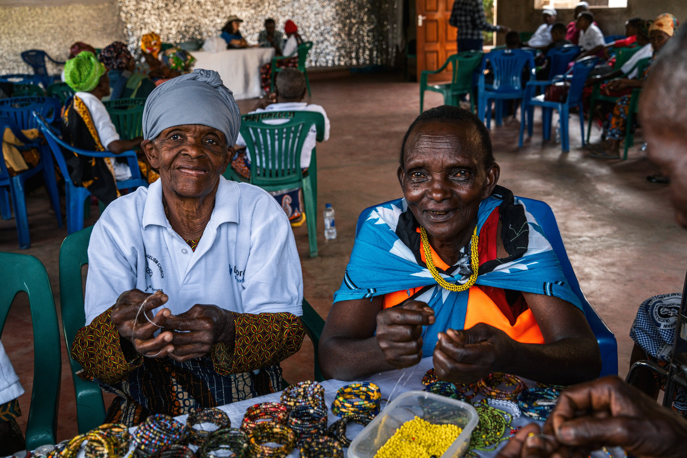 Tanzaniaanse vrouwen zitten aan een tafel en maken kleurrijke sieraden, onderdeel van een dorcas-project dat lokale ambachten en economische ontwikkeling ondersteunt.