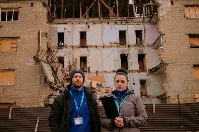 Twee hulpverleners staan voor een beschadigd gebouw in Oekraïne, betrokken bij wederopbouw en humanitaire hulp na de verwoestingen.