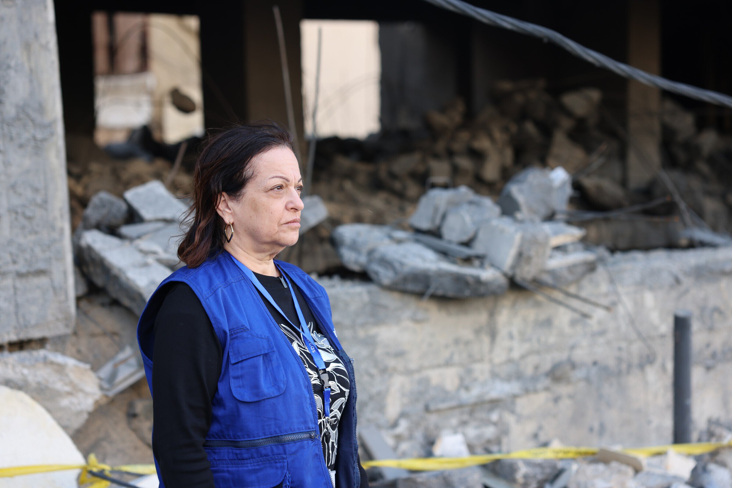 Vrouw in een blauwe vest staat voor de ruïnes en schade na een ramp in Libanon, met zicht op verbrande en ingestorte gebouwen op de achtergrond.