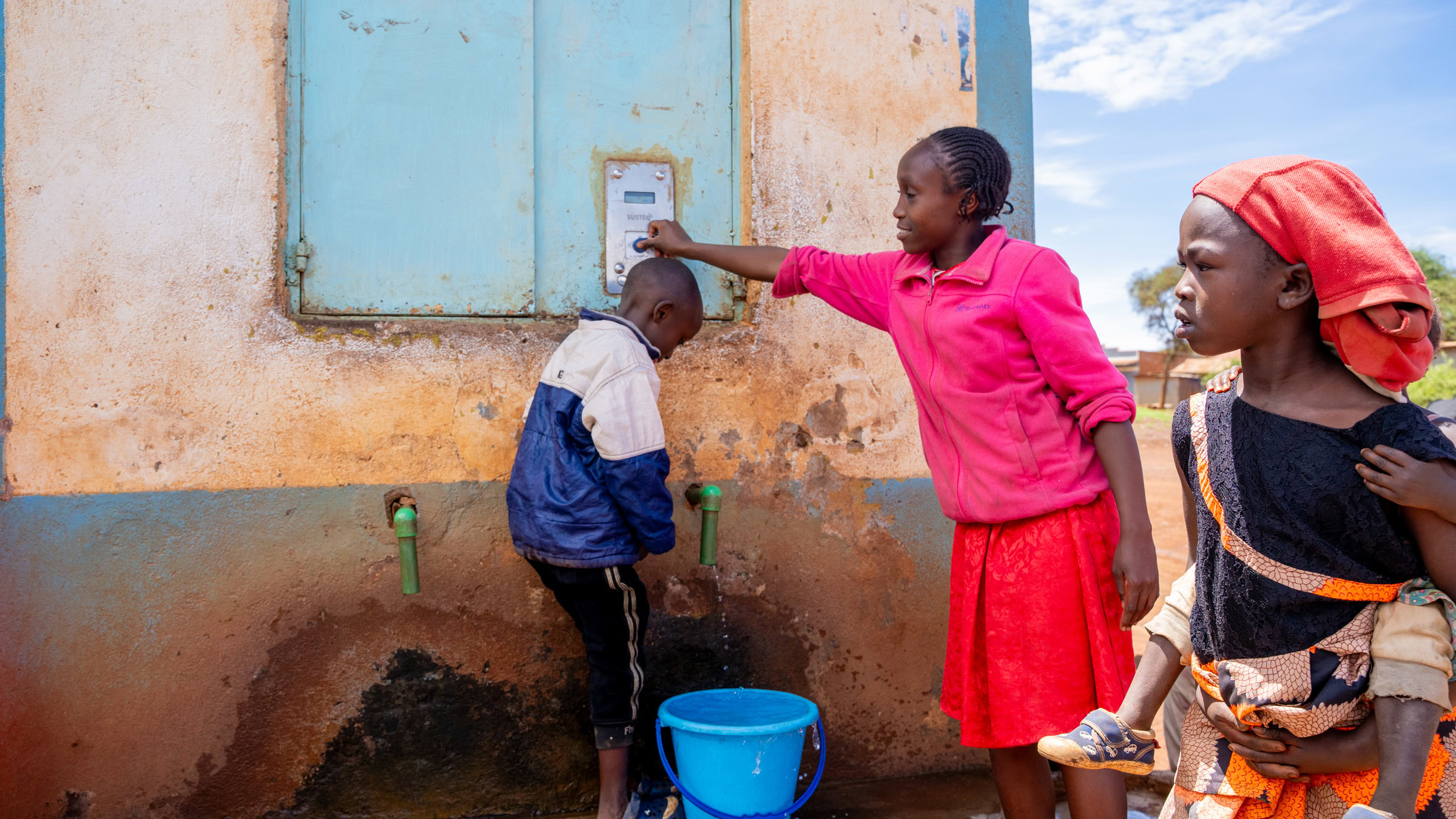 Een vrouw helpt een kind met water halen bij een waterpunt buiten, terwijl een ander kind toekijkt.