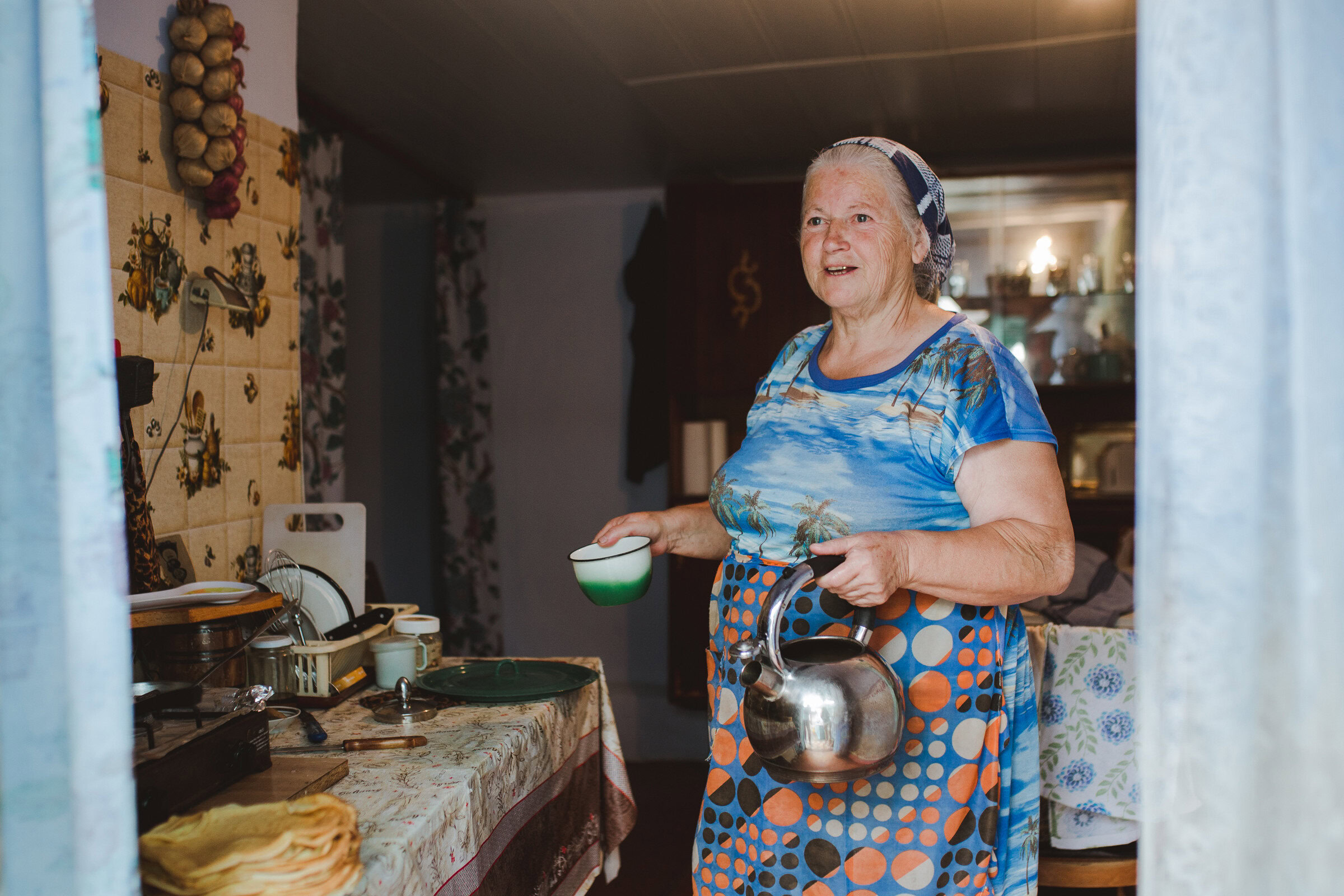 Oudere vrouw in keuken met pollepel en waterkoker, genietend van koken in haar huis, met traditionele decoraties op de muur, passend bij de thema's van hulp en gemeenschap in Moldavië.