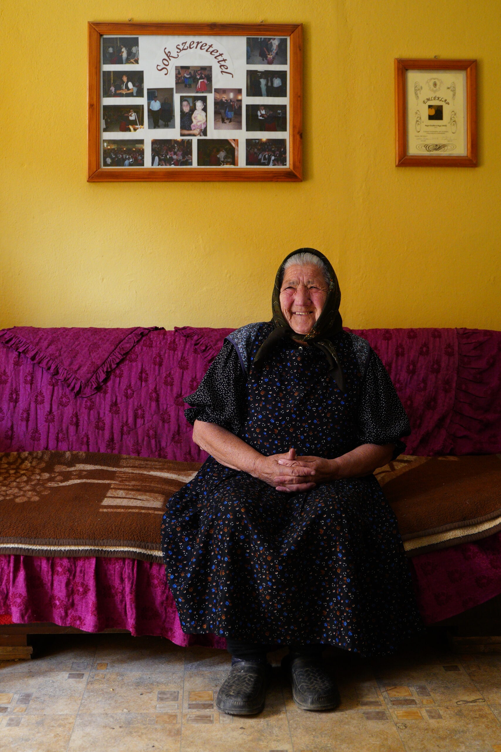 Een oudere vrouw zit lachend op een bank in haar woonkamer, omringd door kleurrijke muren en fotolijsten.