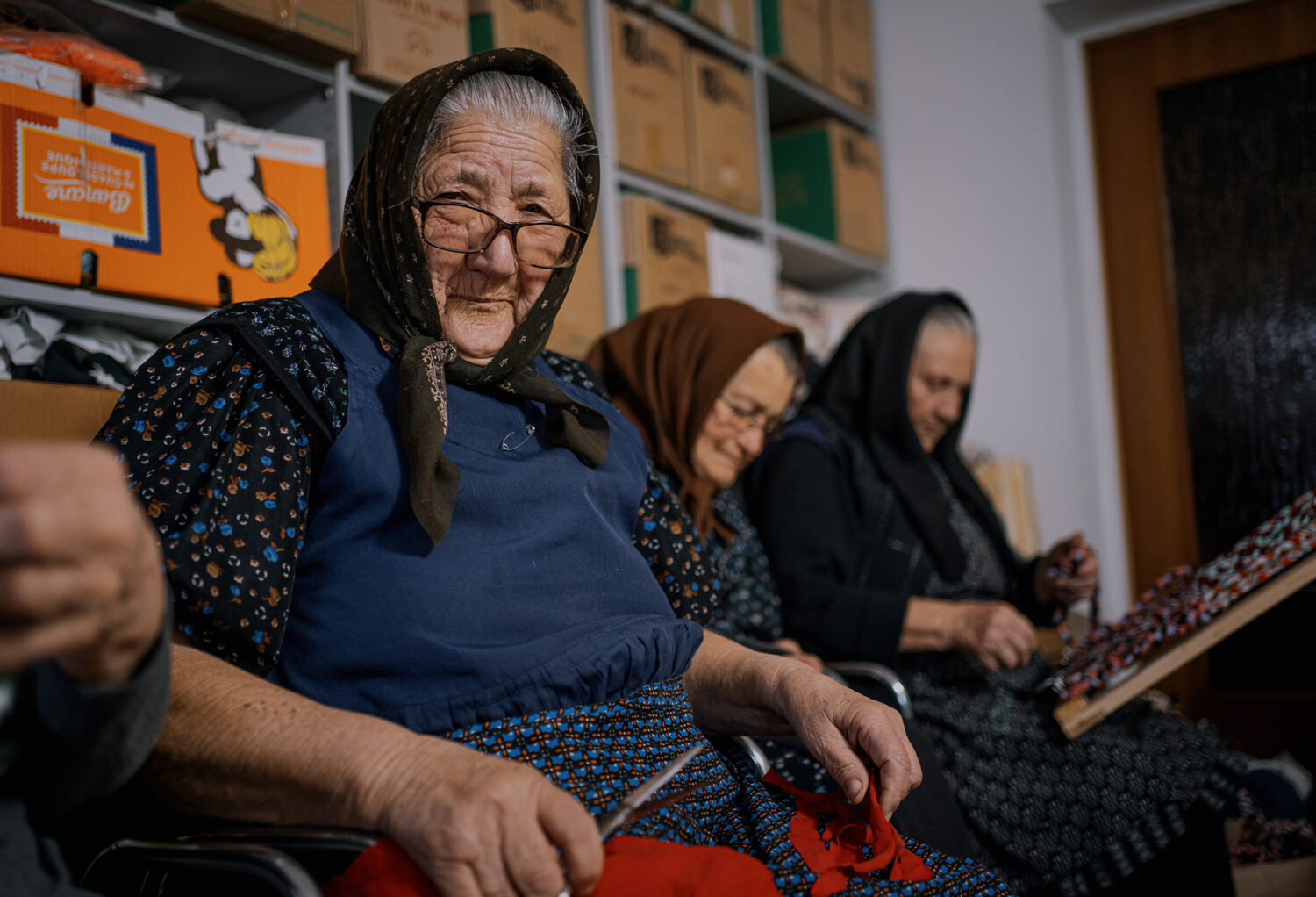 Een groep oudere vrouwen zit samen in een ruimte, mogelijk een winkel of werkplaats, en werken aan creatieve projecten of handwerk.
