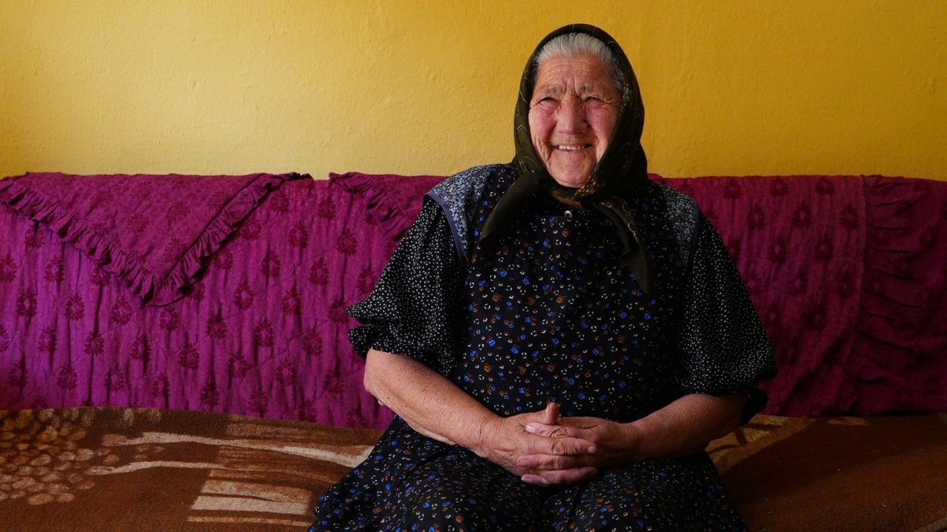 Een oudere vrouw met een glimlach zit op een bank, haar handen gevouwen in haar schoot, in een huis met een gele muur en een paarse bank.