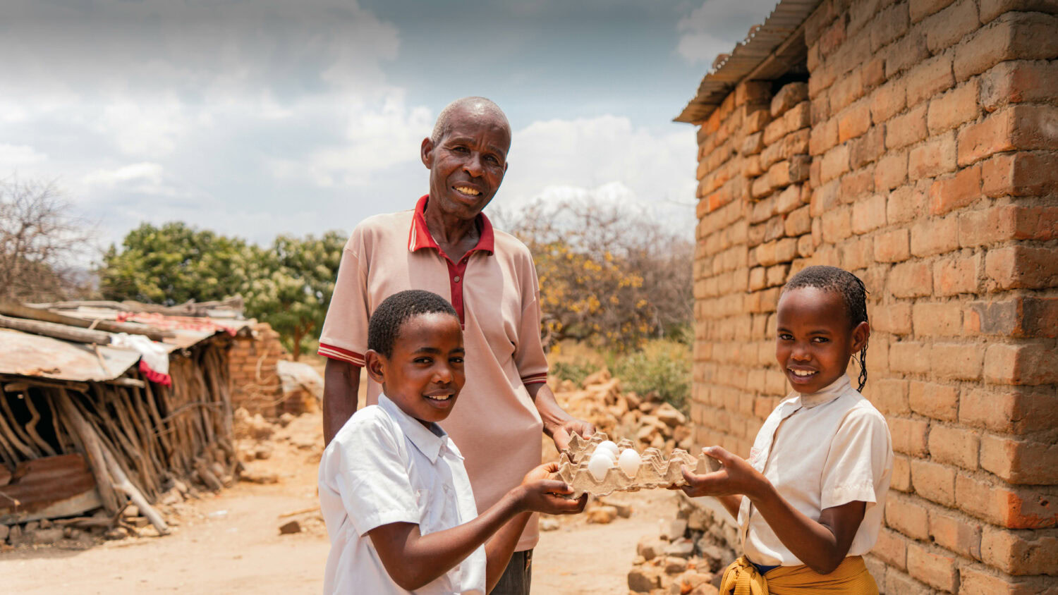 Foto met een man en twee kinderen in Tanzania die kippen hebben gekregen en kunnen leven van de opbrengst. Kinderen en man lachen naar de camera. Foto gemaakt in Tanzania