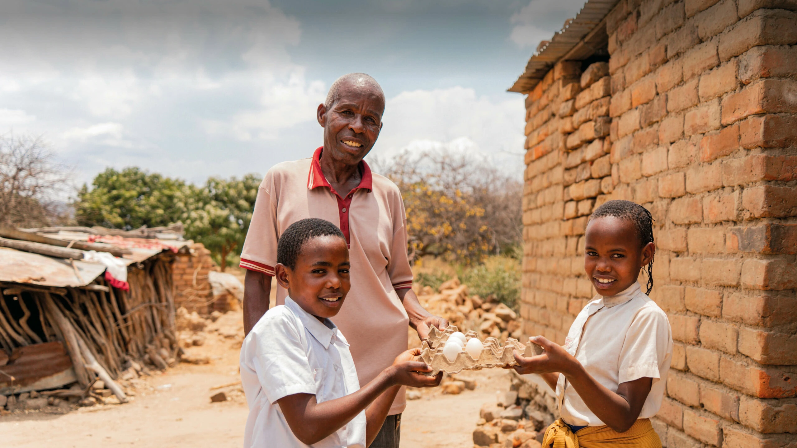 Foto met een man en twee kinderen in Tanzania die kippen hebben gekregen en kunnen leven van de opbrengst. Kinderen en man lachen naar de camera. Foto gemaakt in Tanzania