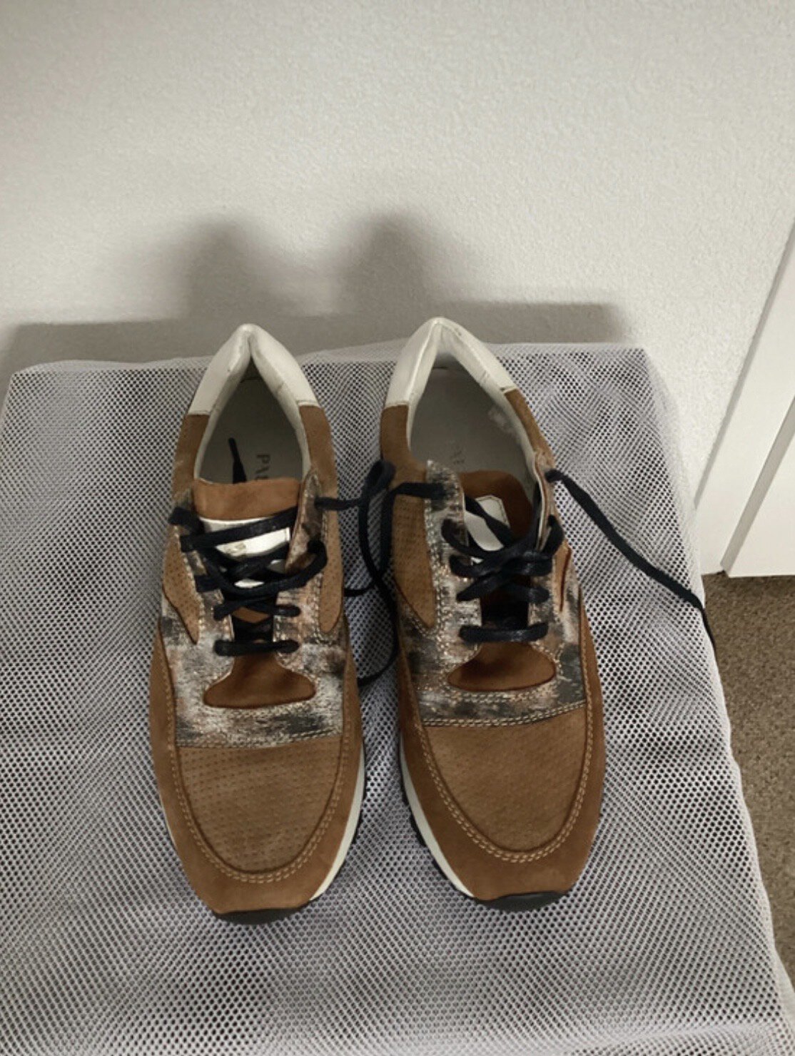 Lage sneakers in bruin en grijs met zwarte veters, geplaatst op een grijs textieloppervlak tegen een witte muur.
