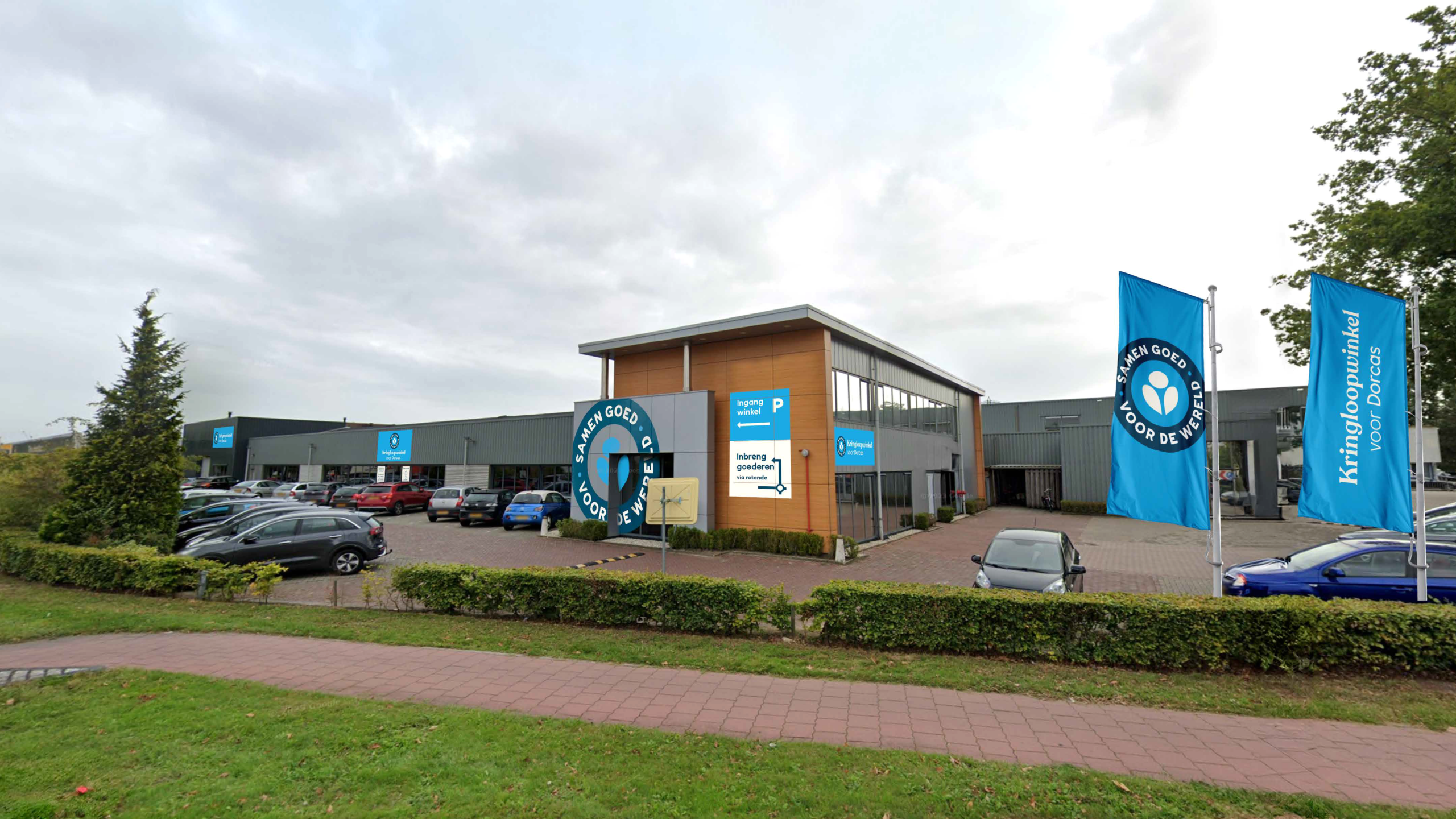 Een overzicht van een kringloopwinkel met parkeerplaats en blauwe vlaggen, gelegen in een groene omgeving, geschikt voor hergebruik en duurzame consumptie.