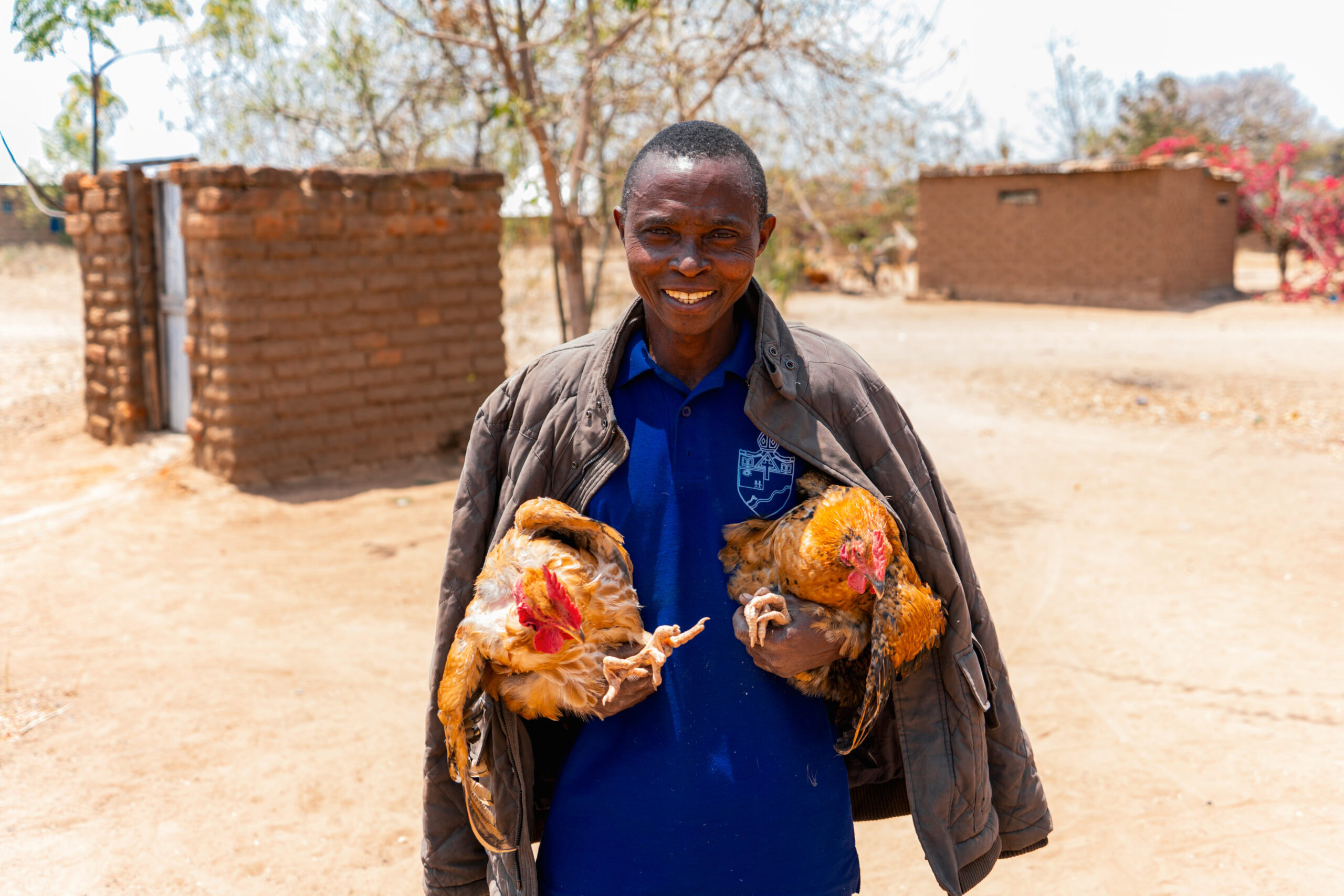 Een man in Tanzania houdt twee kippen, lachend en trots, in een dorpsomgeving met eenvoudige huisjes en droge grond.
