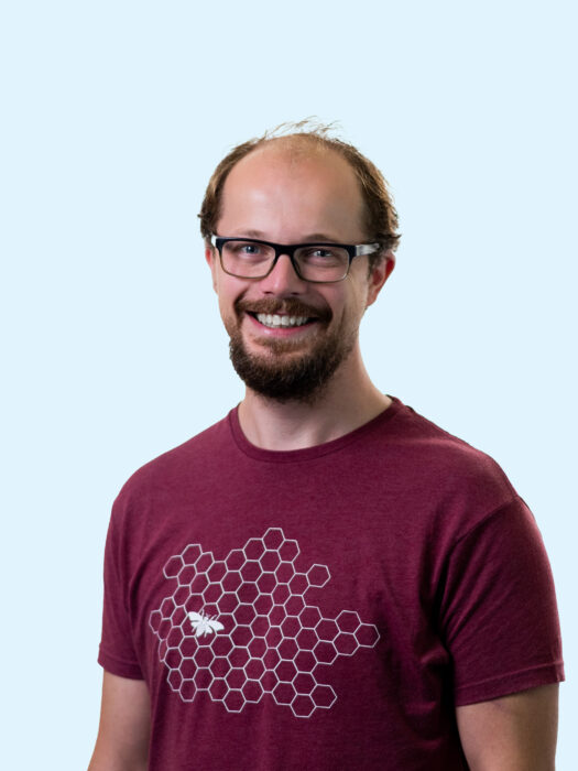 Een man met een bril en een baard, lachend, draagt een bordeauxrood T-shirt met een hexagonale patroon en een bij.