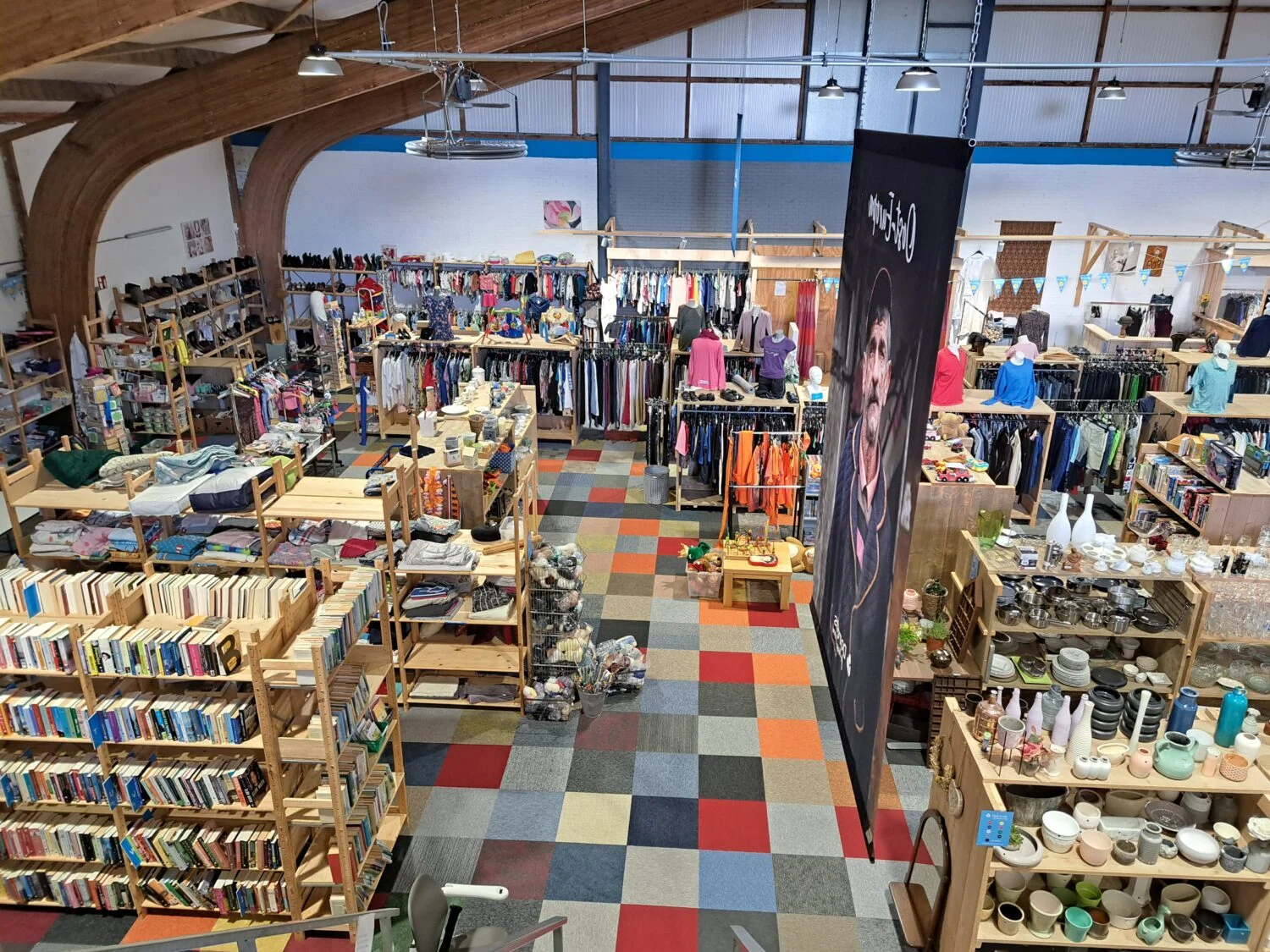 Interieur van een tweedehandswinkel met boeken, kleding en huishoudartikelen, georganiseerd op houten rekken en planken, met kleurrijke vloerbedekking en een open, lichte ruimte.