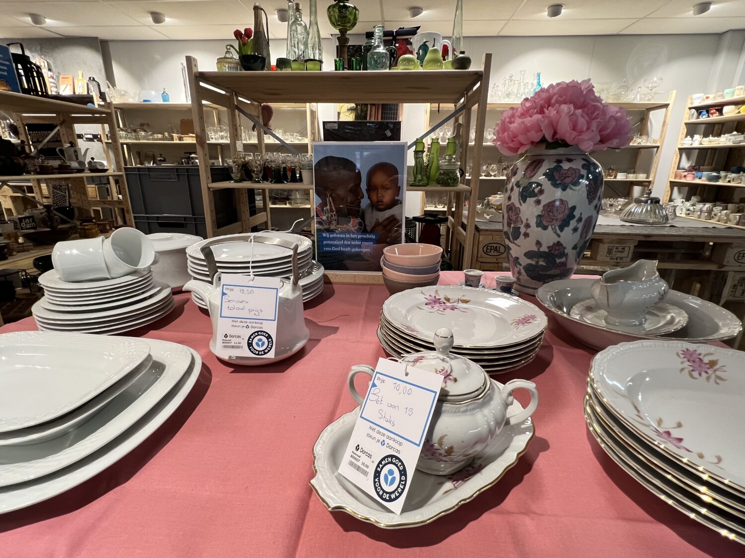 Een uitgebreide collectie porseleinen servies met bloemenmotief, inclusief borden, kopjes en een theepot, gepresenteerd op een tafel met een roze tafelkleed in een winkel.