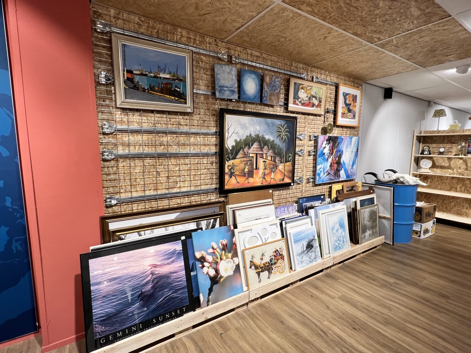 Een collectie van diverse kunstwerken en schilderijen, uitgestald op een muur en op de vloer, in een gezellige galerie met houten vloer en creatieve sfeer.