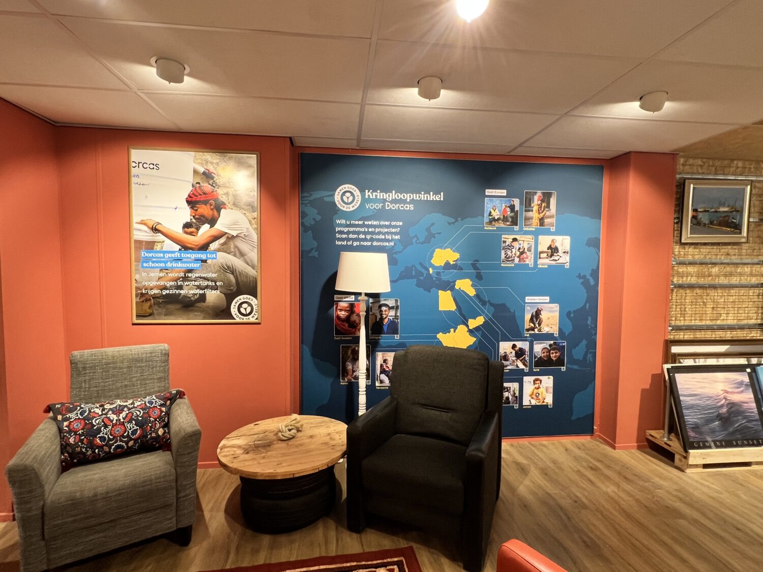 Gezellige zithoek met twee stoelen, een ronde tafel en een lamp, gelegen in een kantoorruimte van Dorcas met een wereldkaart en foto's op de muur.