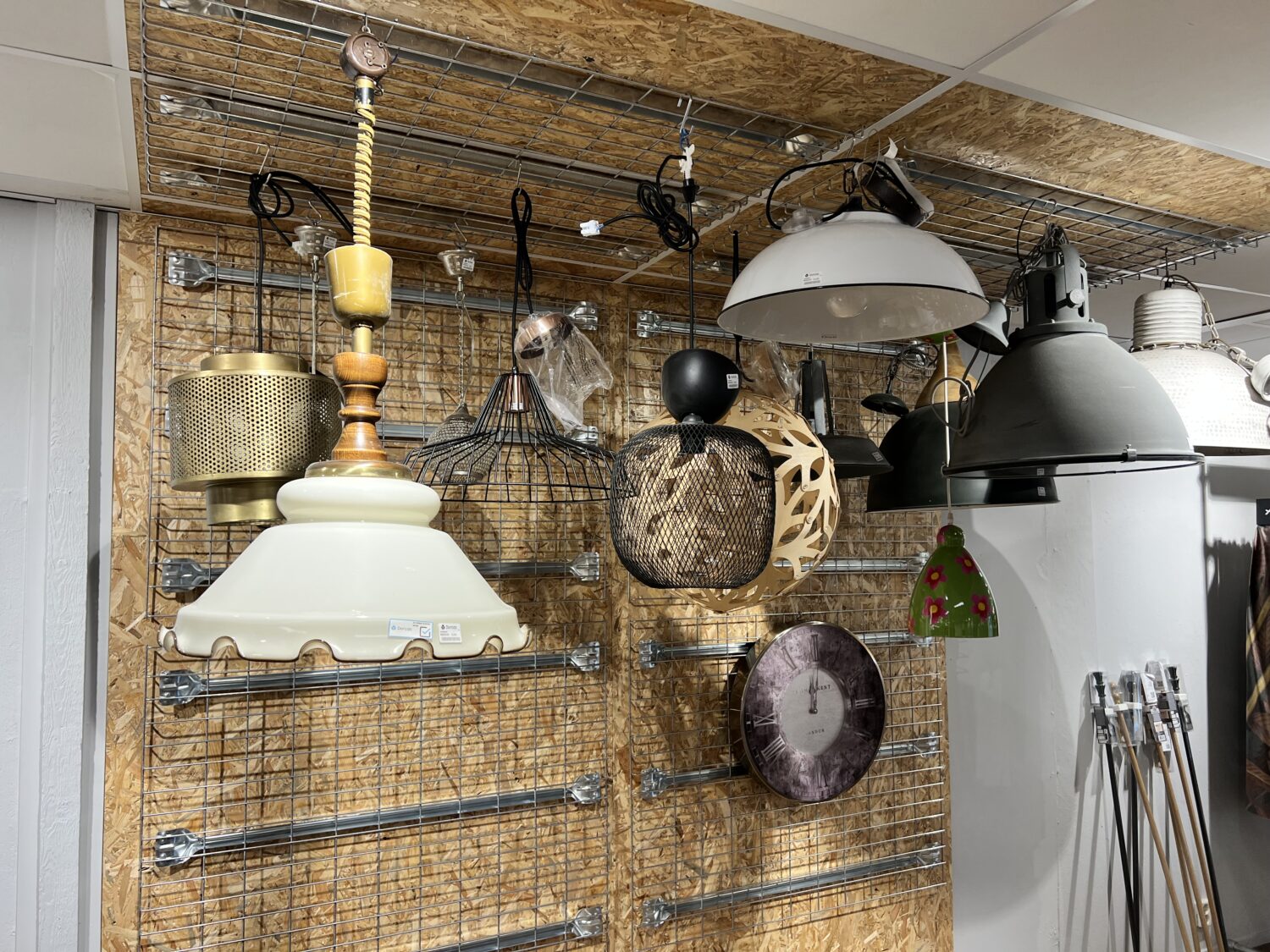 Diverse lampen en lampenkappen hangen aan een metalen rooster tegen een houten wand, geschikt voor interieurdecoratie of verlichtingswinkel.