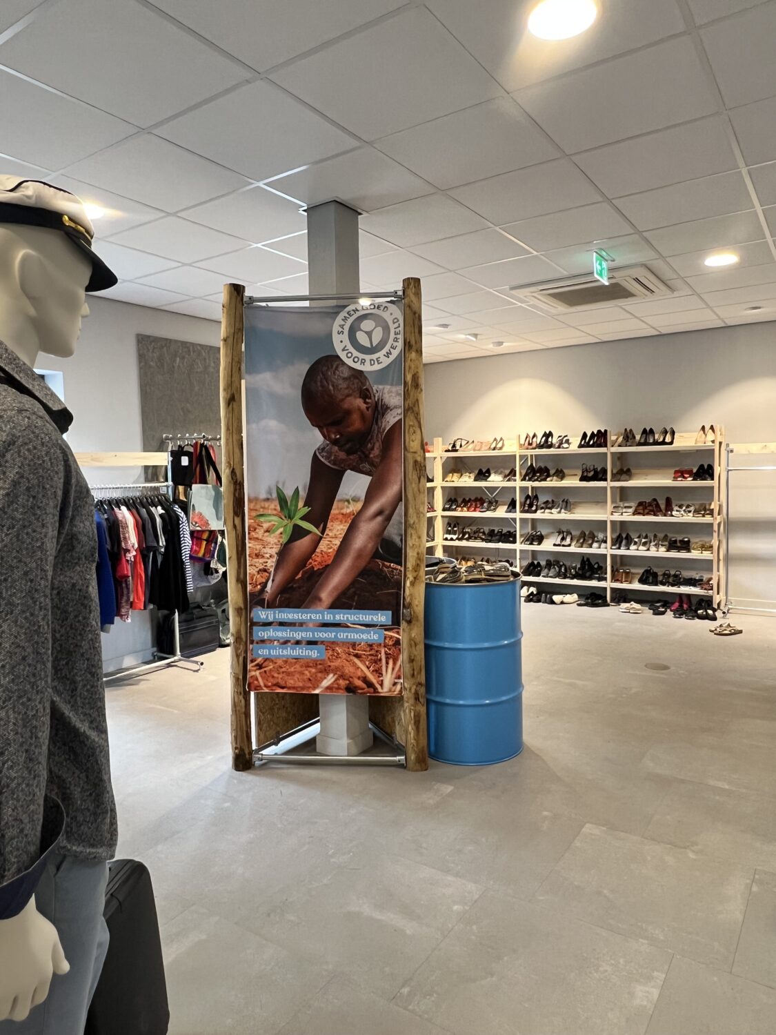 Binnen in een winkel van Dorcas, met een display en schoenen op de achtergrond, wordt een poster getoond die het sociale project van Dorcas promoot en mensen aanmoedigt om te helpen.