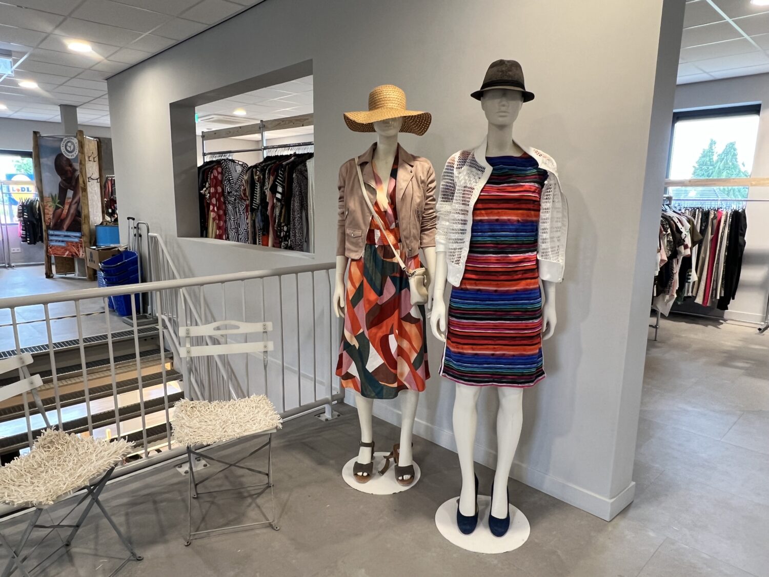 Twee mannequins in een kledingwinkel dragen zomerse jurken en hoeden, met andere kledingrekken op de achtergrond, in een moderne winkelomgeving.