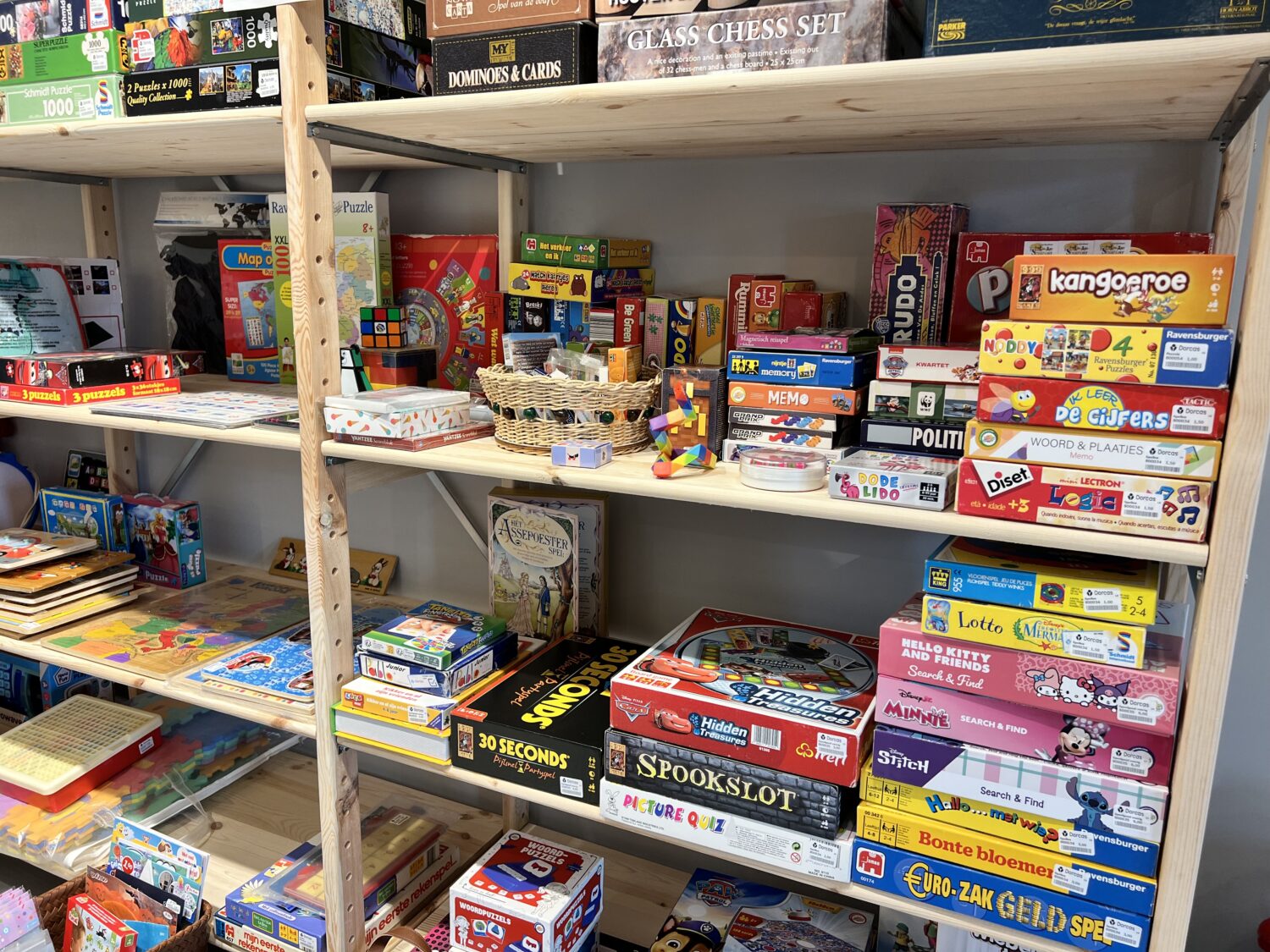 Een overzicht van diverse speelgoed- en bordspellen op houten rekken, inclusief bekende merken zoals Monopoly, Catan en 30 Seconds, geschikt voor kinderen en families.