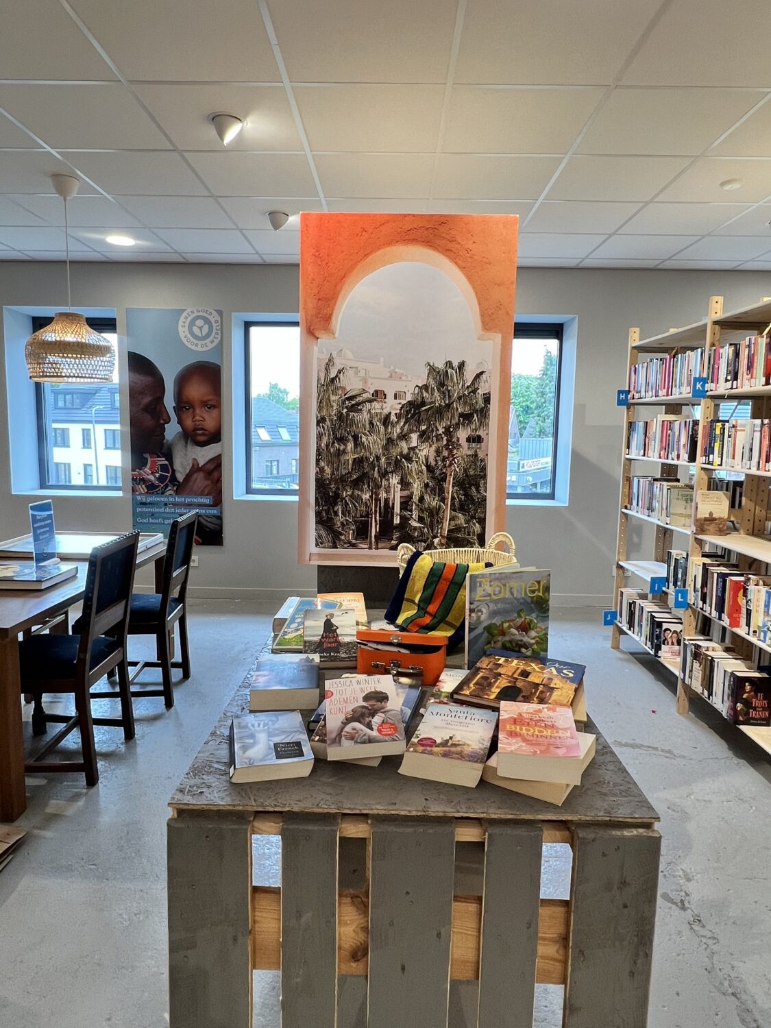 Interieur van een boekwinkel met een tafel vol boeken, een kunstwerk aan de muur, en boekenplanken op de achtergrond, met grote ramen die natuurlijk licht binnenlaten.