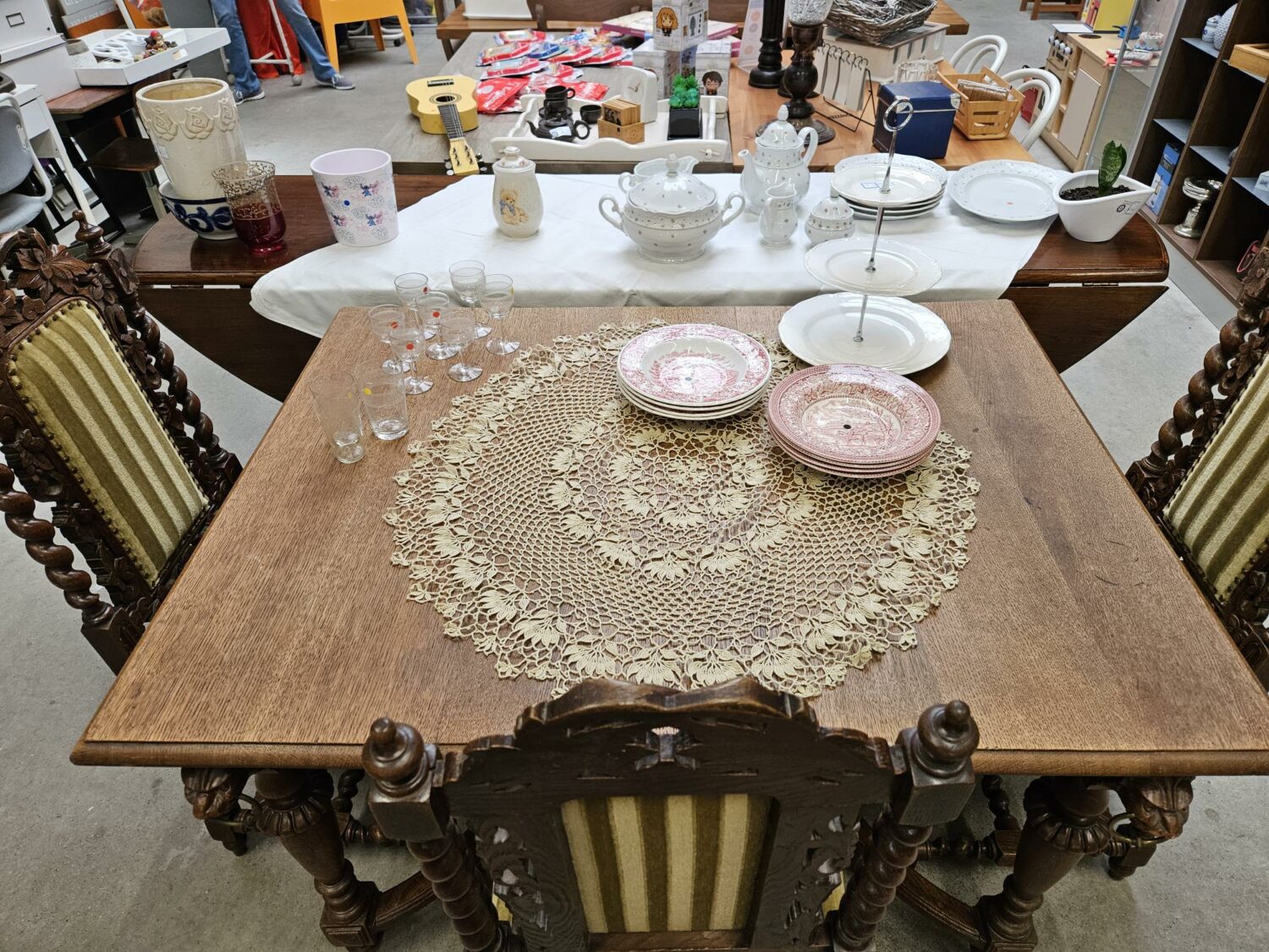 Een houten eettafel met een kanten tafelkleed en servies, omringd door antieke stoelen, in een tweedehandswinkel of antiekzaak, klaar voor verkoop of gebruik.