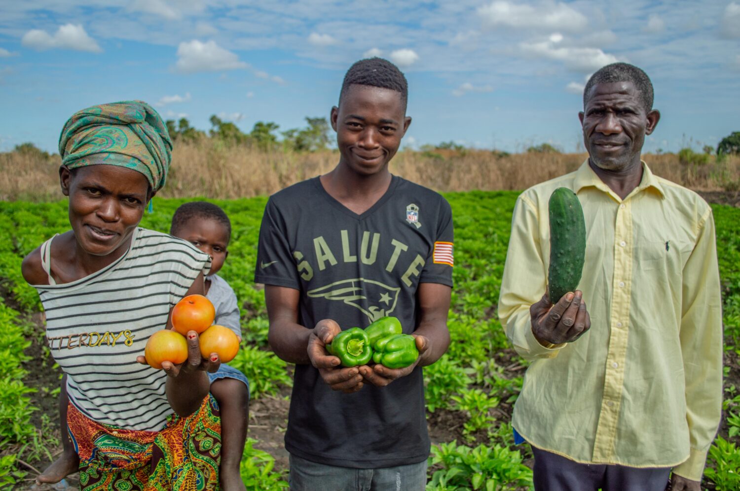 Een familie in Afrika toont groenten en fruit die ze verbouwen, met een veld en blauwe lucht op de achtergrond.