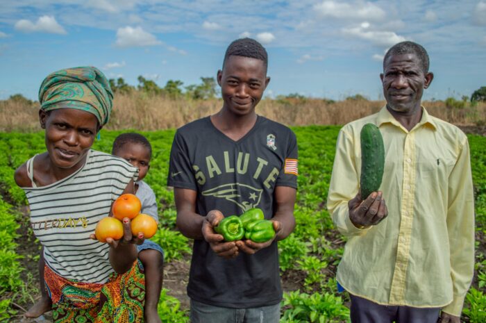 Een familie in Afrika toont groenten en fruit die ze verbouwen, met een veld en blauwe lucht op de achtergrond.