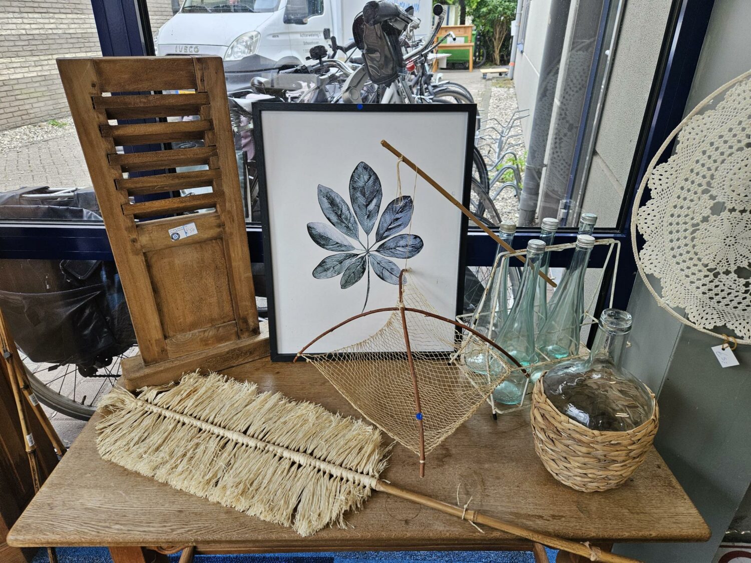 Een collectie vintage decoratie en kunst op een houten tafel, inclusief een houten stoel, een schilderij van bladeren, glazen flessen, een mand met een glazen pot, en een veer met een net, symbolise-.