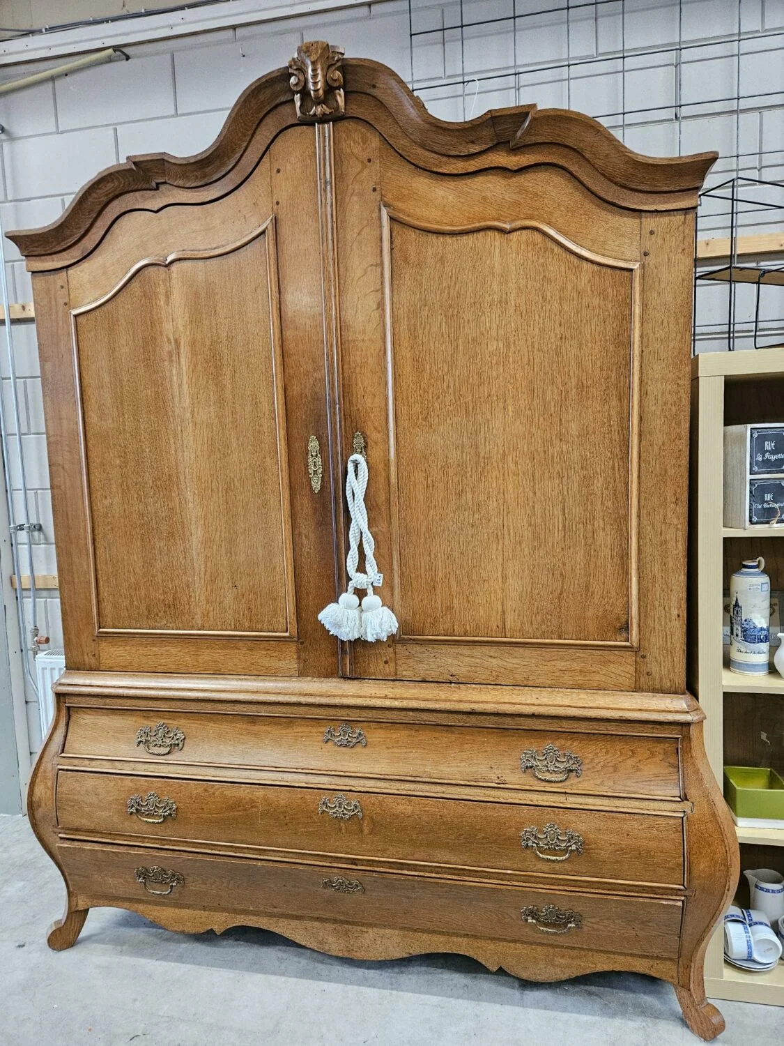Antieke houten kast met sierlijke details en een witte tassel, geschikt voor opslag en decoratie, getoond in een winkel of opslagruimte.