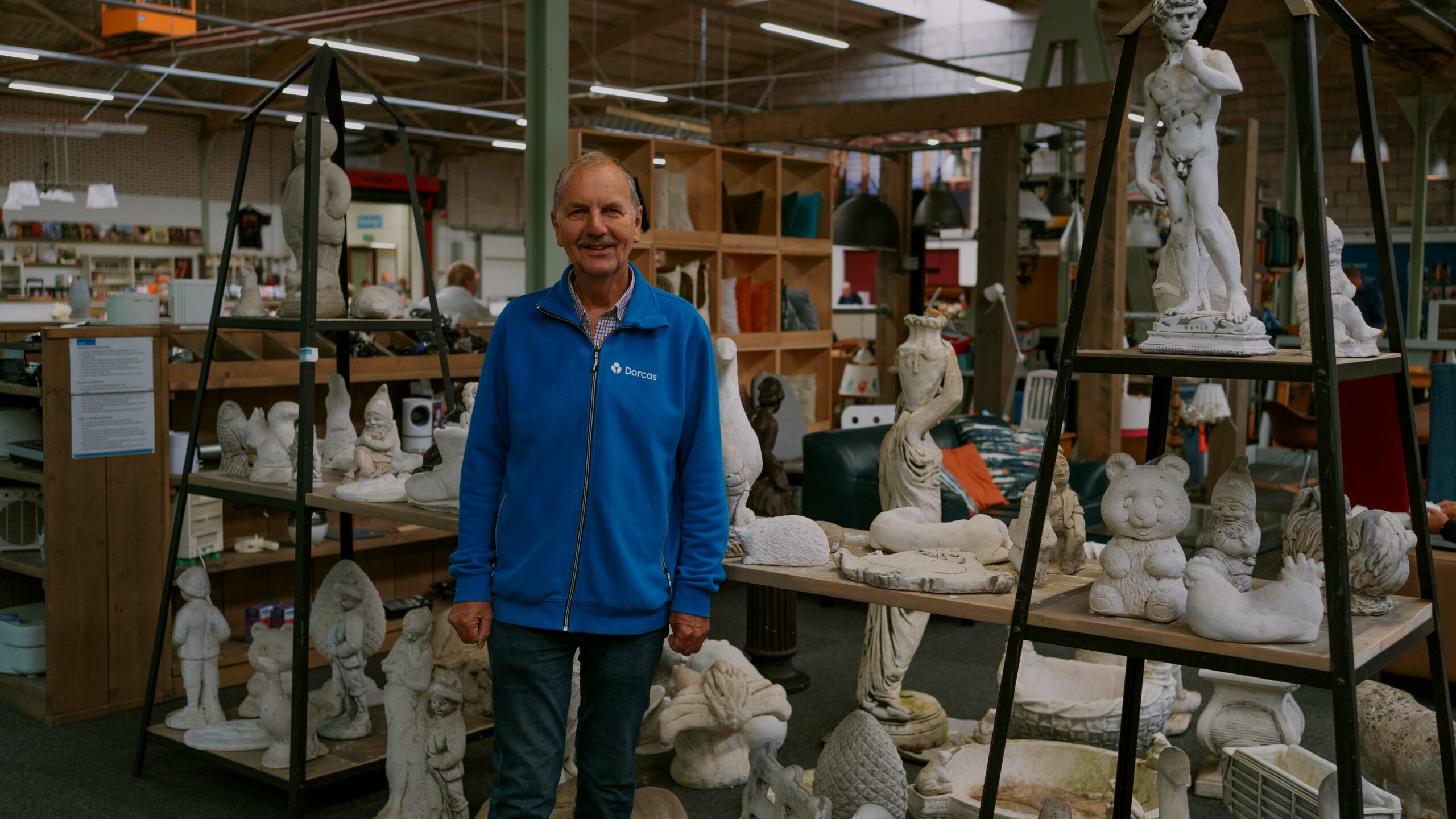 Man in blauwe jas staat tussen sculpturen en kunstwerken in een atelier of winkel, symboliseert steun en creativiteit voor humanitaire projecten van Dorcas.