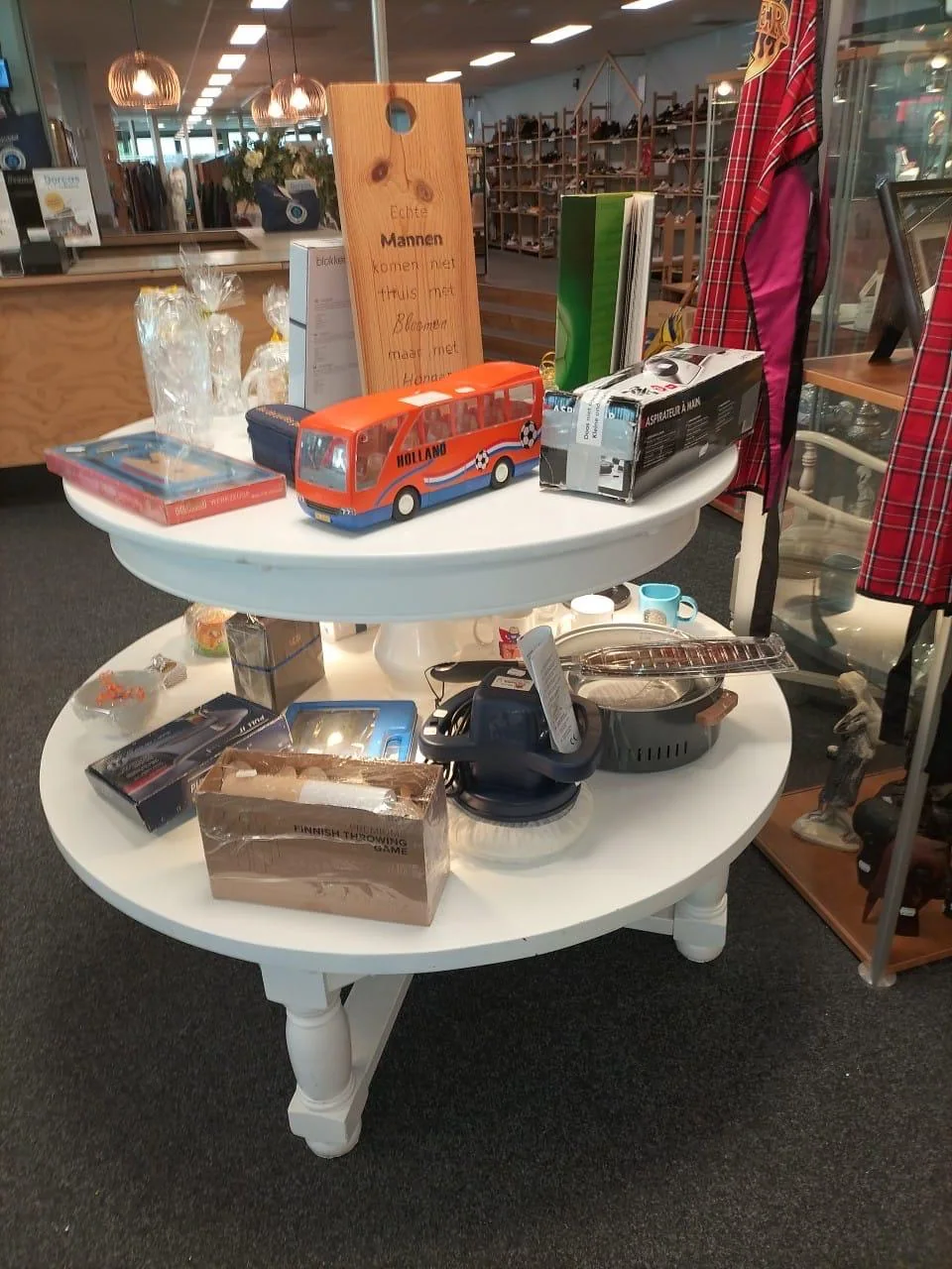 Een ronde witte tafel met speelgoedauto's, boeken, een houten bord, en huishoudelijke artikelen in een tweedehandswinkel, met winkelinterieur op de achtergrond.