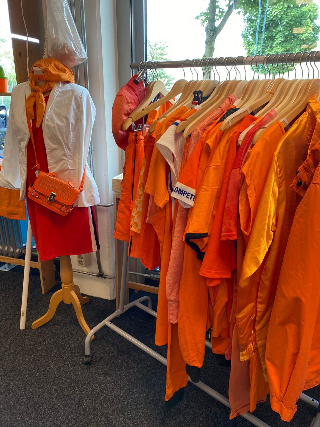 Zomerkleding en oranje accessoires hangen aan een kledingrek en een mannequin met een oranje tas, binnen in een winkel met grote ramen en groene bomen buiten.