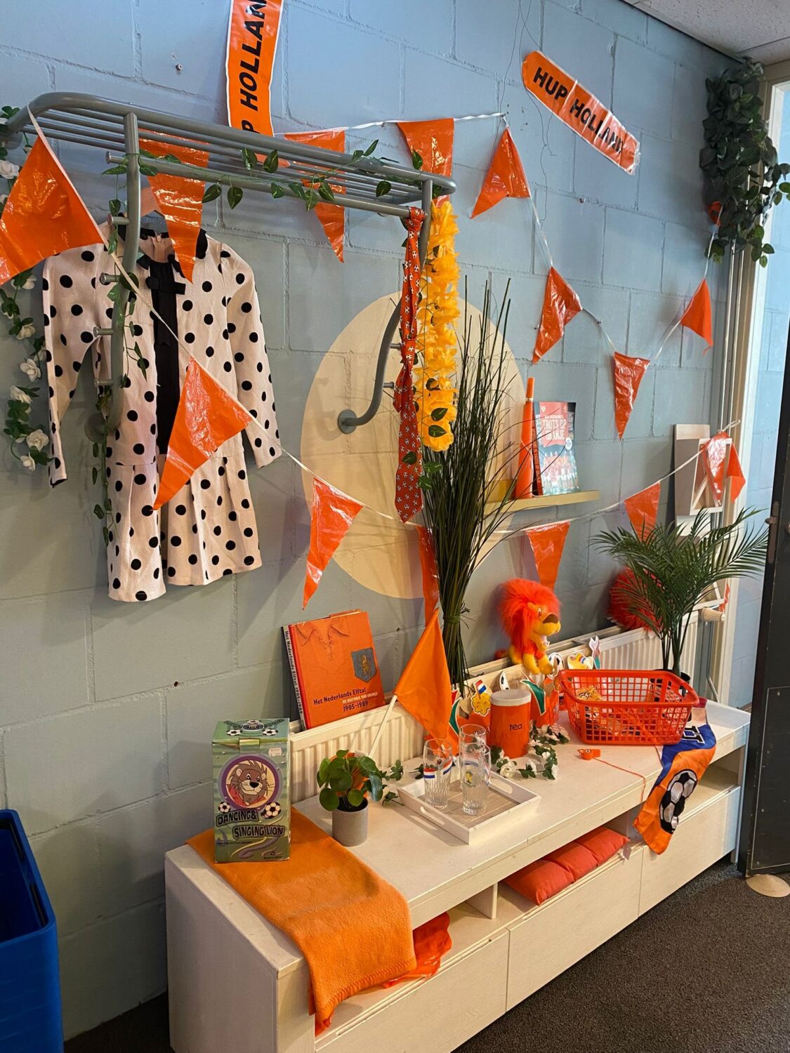 Een vrolijk ingerichte winkelruimte met oranje feestdecoraties, een kledingrek met een polka dot jas, en diverse accessoires en planten op een witte bank, klaar voor een feestelijke gebeurtenis.