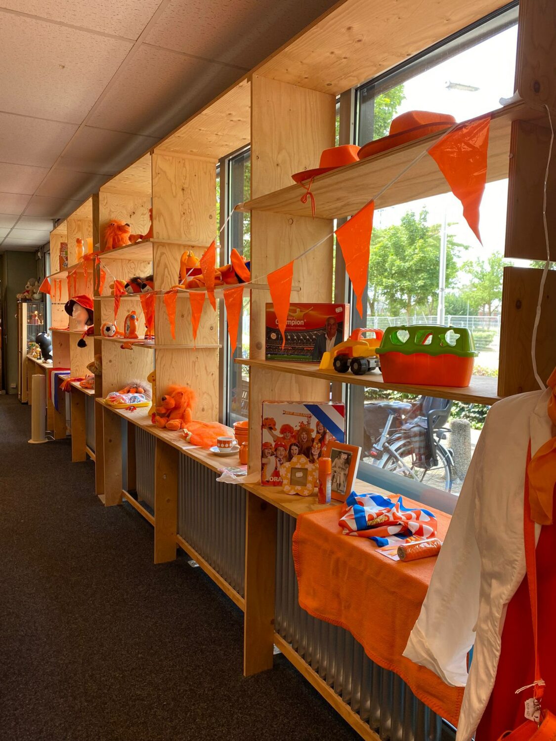 Een interieur met houten wandpanelen en grote ramen, versierd met oranje vlaggetjes en speelgoed, mogelijk voor een feest of evenement bij Dorcas.