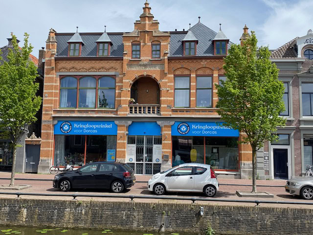 Kringloopwinkel voor Dorcas in een historisch rood bakstenen gebouw met blauwe winkeluitbatingsborden, omgeven door groene bomen en geparkeerde auto's, in een stadscentrum.