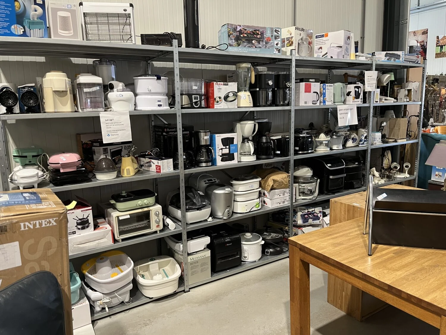 Diverse huishoudelijke apparaten en keukenartikelen op metalen planken in een winkel, met een houten tafel en stoel op de voorgrond, geschikt voor noodhulp en ontwikkelingsprojecten.