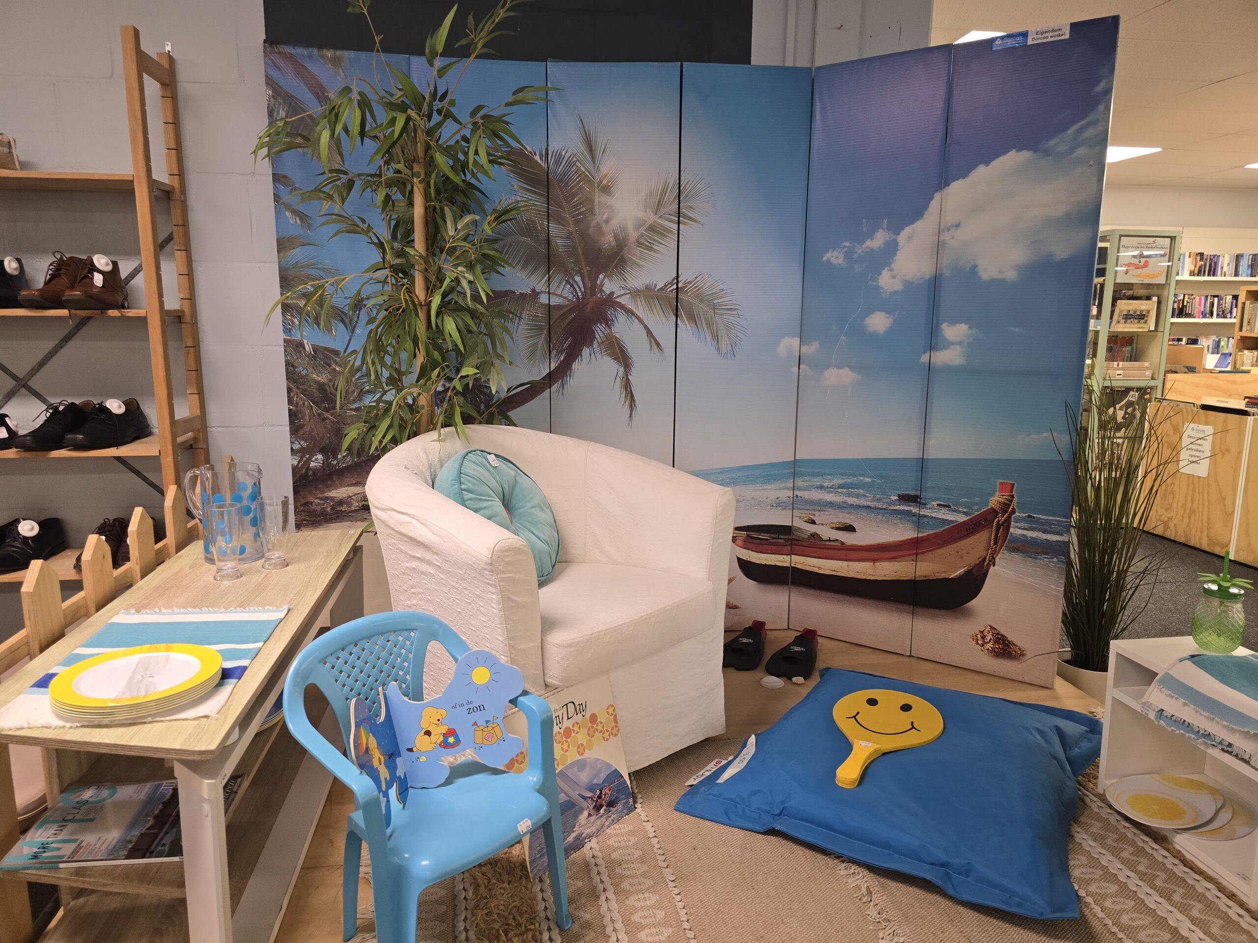 Gezellige leeshoek met strandthema, inclusief een witte fauteuil, een blauwe kinderstoel met zeefiguren, een grote strandfoto als achtergrond, en een blauwe kussen met een smiley, geschikt voor humanj.