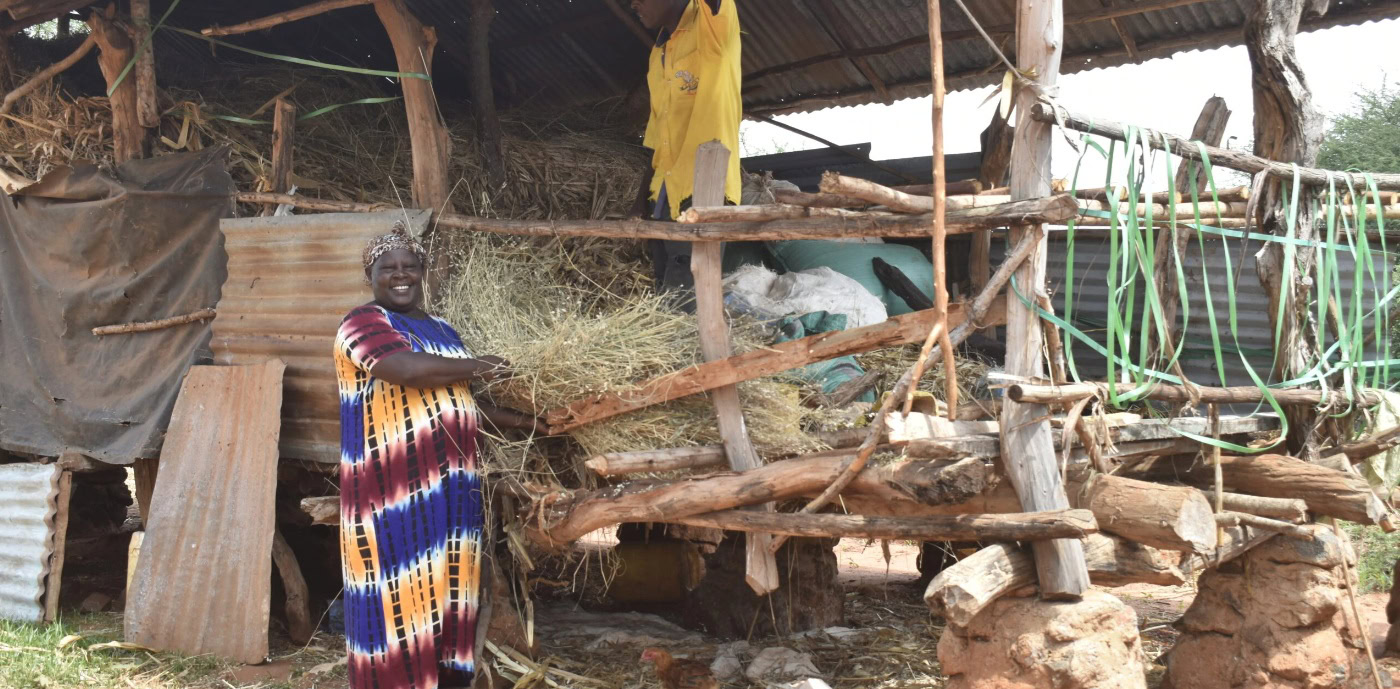 Een vrouw in kleurrijke kleding staat voor een eenvoudige houten hut in Oost-Afrika, symboliserend humanitaire hulp en armoedebestrijding door Dorcas.