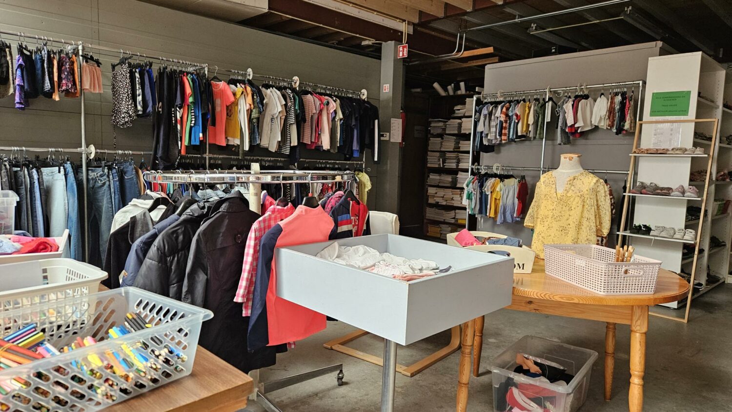 Een tweedehands kledingwinkel met kledingrekken vol diverse kledingstukken, een tafel met accessoires en een mannequin met een gele blouse, gericht op humanitaire hulp en armoedebestrijding.