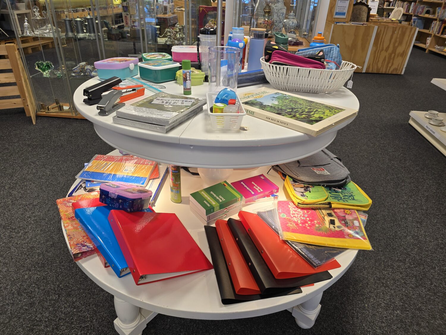 Twee witte ronde tafels met schoolspullen zoals boeken, pennen, notitieboekjes en knutselmaterialen, in een bibliotheek of winkel, symboliseert educatieve hulp en armoedebestrijding.