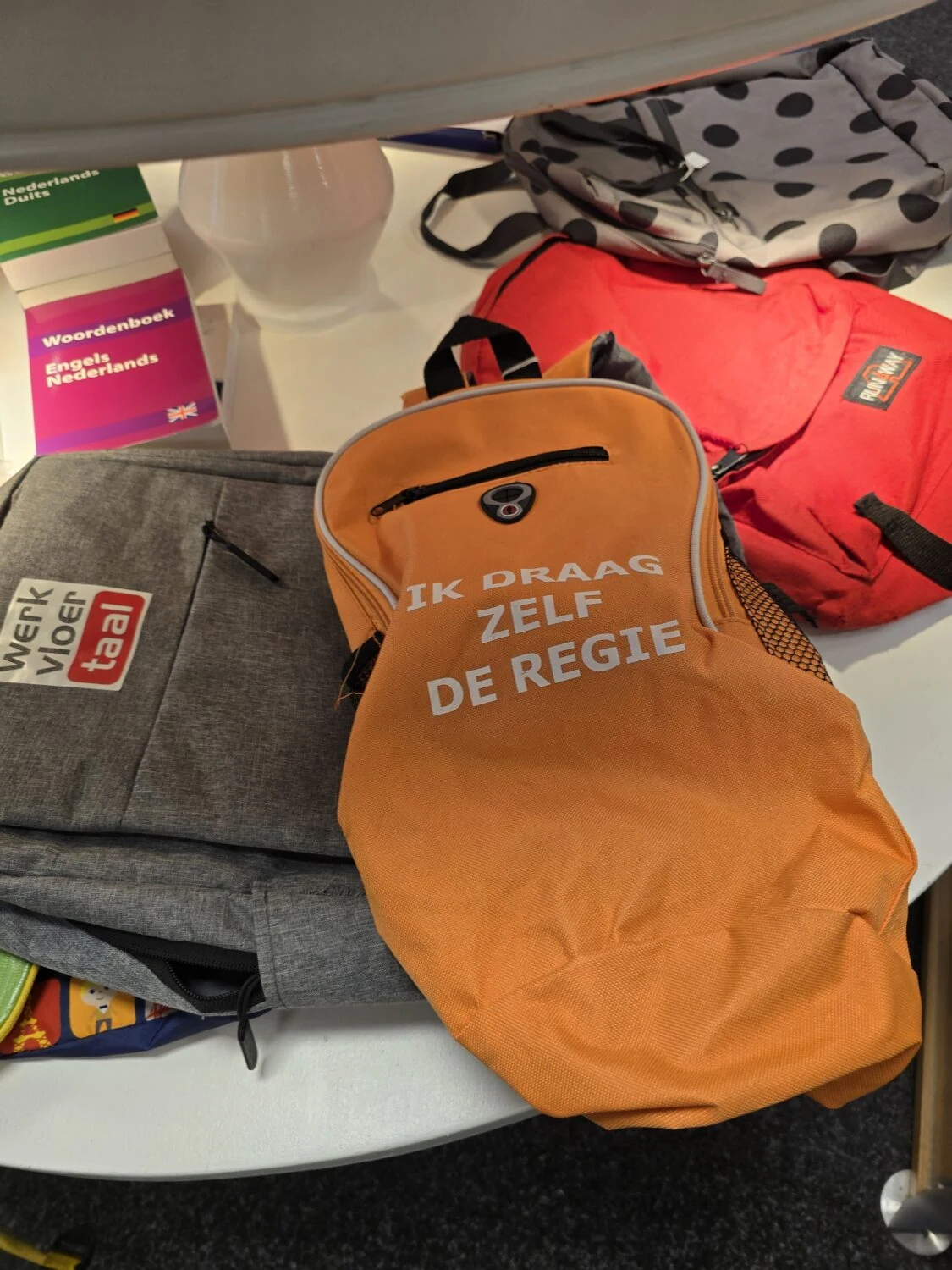 Oranje rugzak met tekst 'Ik draag zelf de regie' op een tafel met andere tassen en boeken, symboliseert empowerment en zelfredzaamheid in humanitaire hulp.