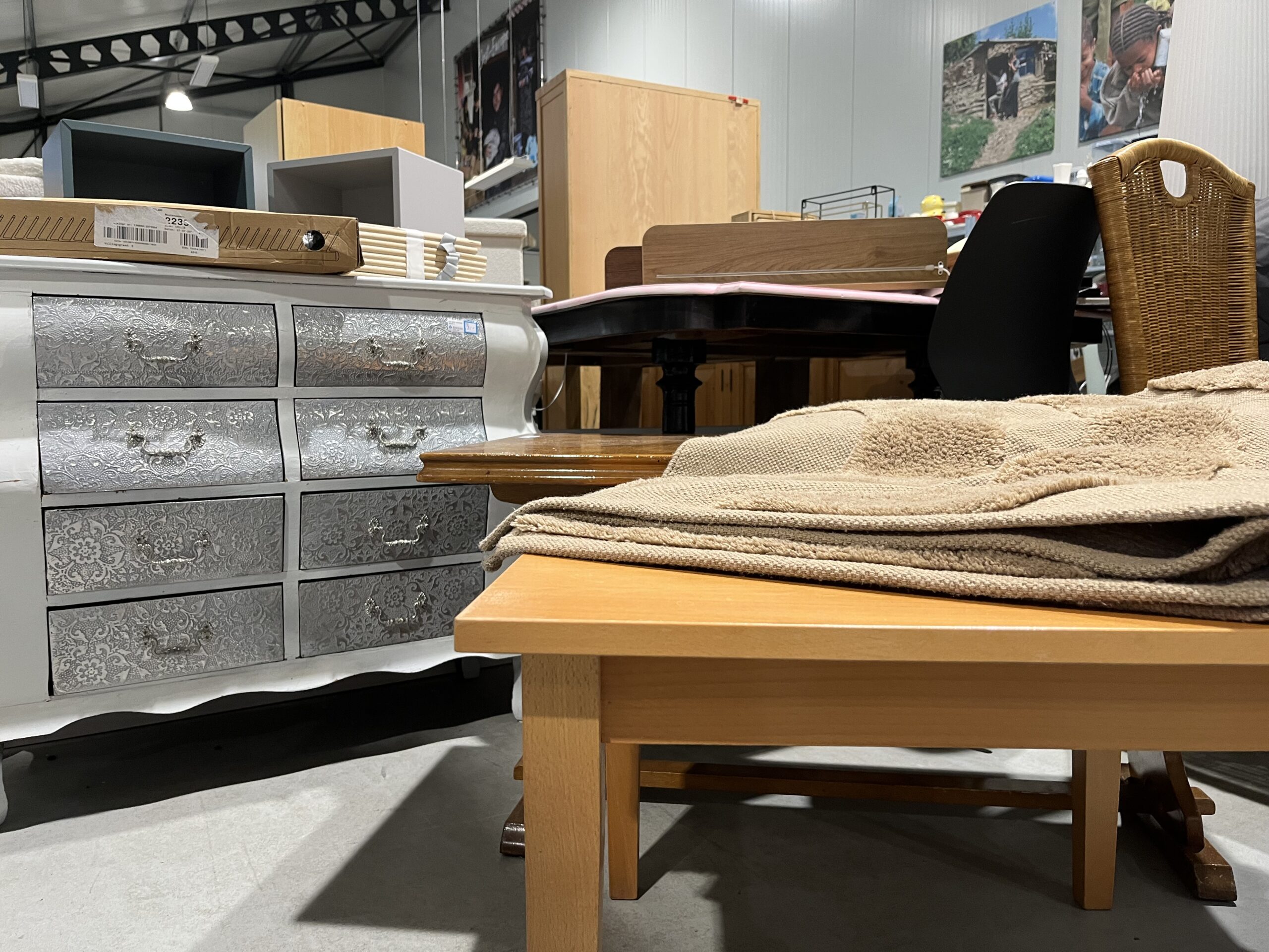 Binnenkijkje in een tweedehandswinkel met diverse meubels zoals een houten tafel, ladenkast en stoelen, passend bij humanitaire hulp en armoedebestrijding.