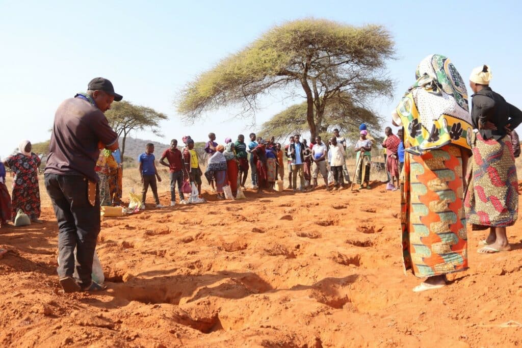 Hulpverleners en lokale bewoners verzamelen zich in een droge, zandige omgeving met enkele bomen, tijdens een humanitaire hulpactie in Oost-Afrika, gericht op armoedebestrijding en noodhulp.
