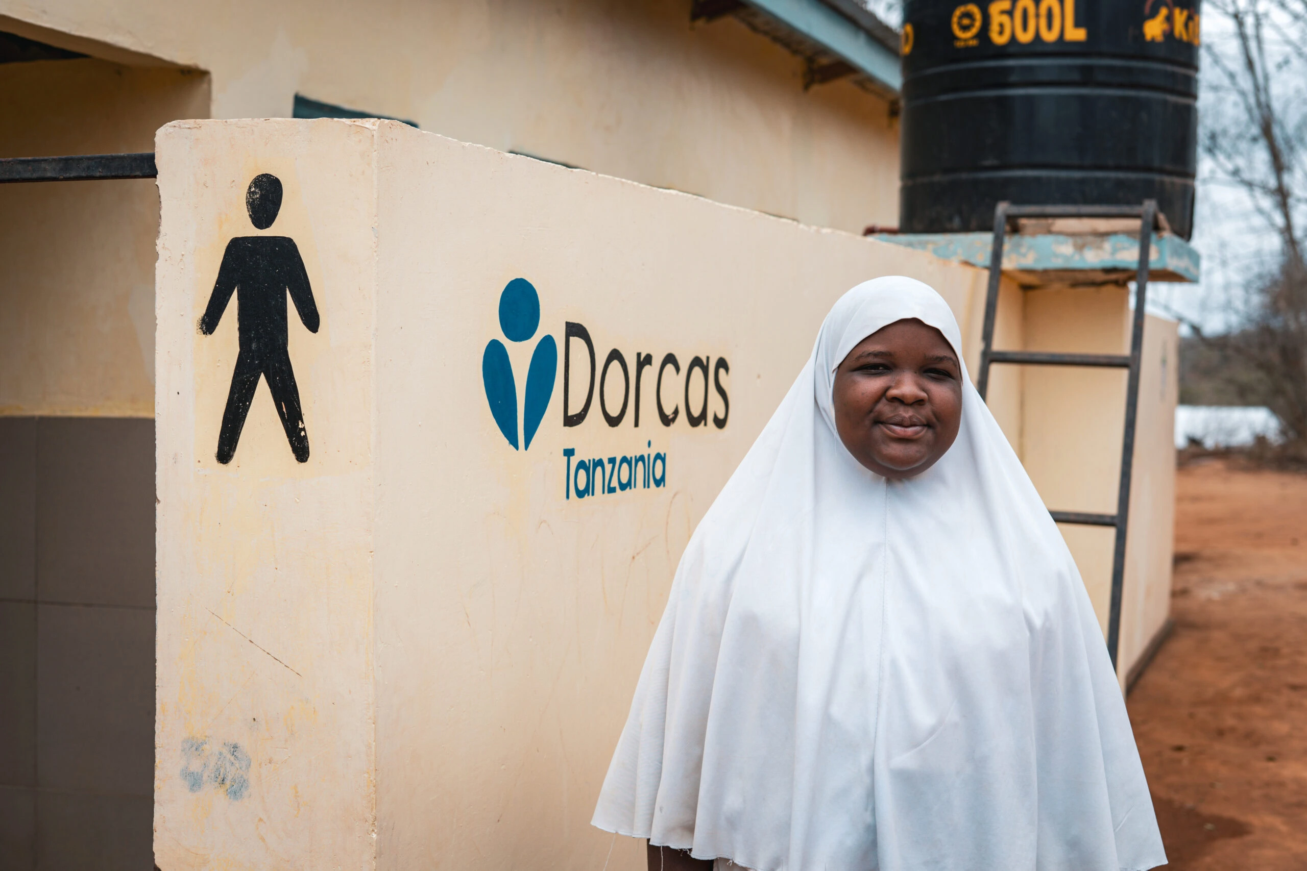 Vrouw in witte hoofddoek voor waterinstallatie met Dorcas Tanzania logo, symboliseert humanitaire hulp en armoedebestrijding in Oost-Afrika.