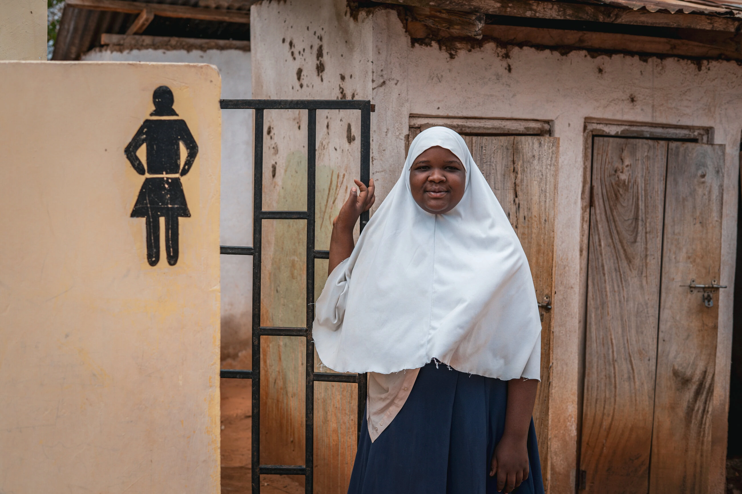 Een jonge vrouw in een hoofddoek staat naast een sanitaire voorziening, symboliseert toegang tot hygiëne en sanitaire voorzieningen in ontwikkelingslanden, belangrijk voor gezondheid.