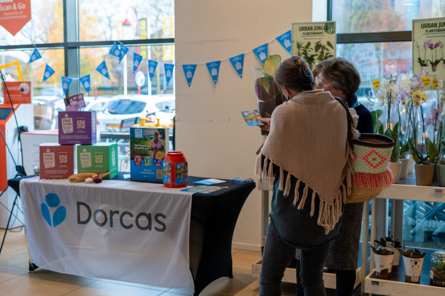Twee vrouwen praten bij de Dorcas-informatietafel met informatie over humanitaire hulp en armoedebestrijding, binnen een winkel met planten en decoraties.