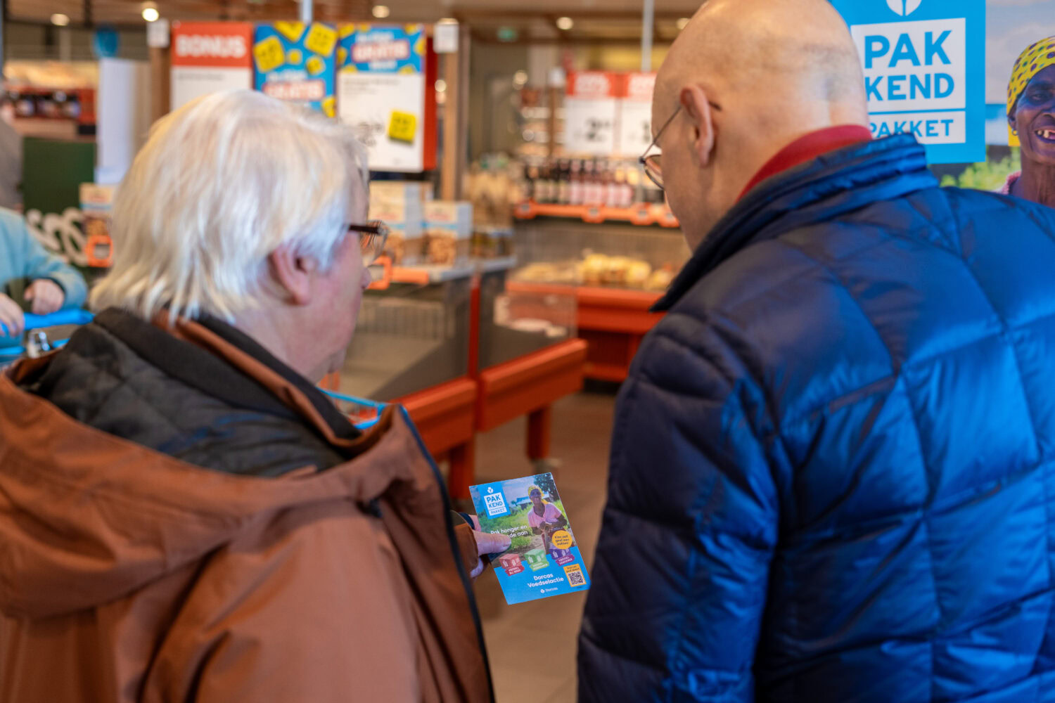 Twee hulpverleners bespreken een flyer in een supermarkt, symboliseert humanitaire hulp en armoedebestrijding, passend bij de missie van Dorcas.