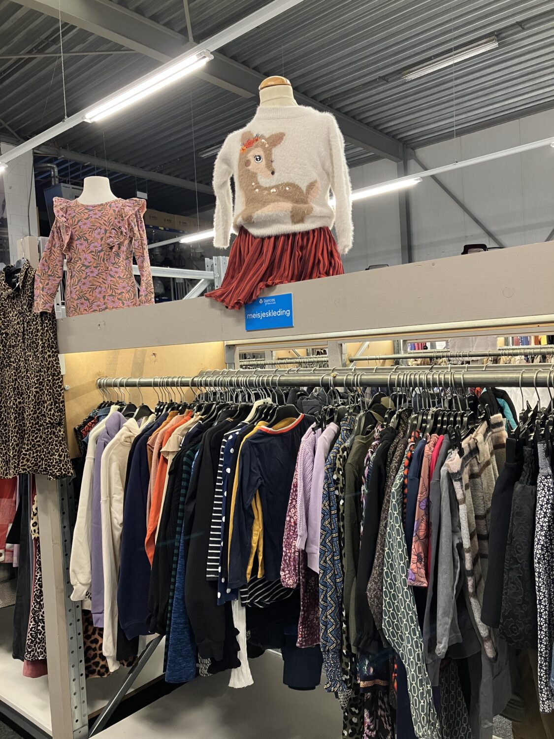 Tweedehands kledingwinkel met mannequins en kledingrekken vol diverse kledingstukken, gericht op humanitaire hulp en armoedebestrijding in ontwikkelingslanden.